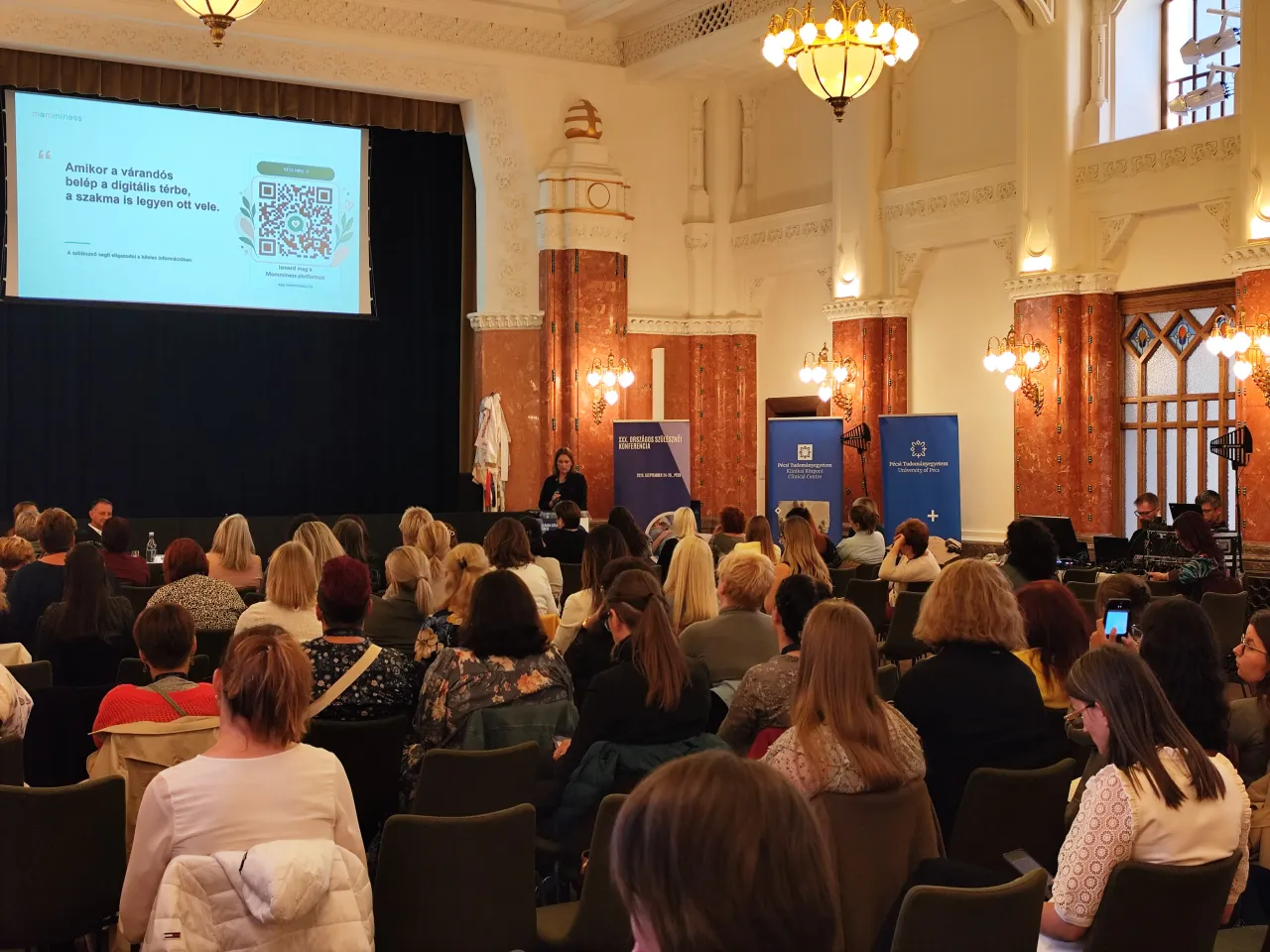 Zsebe-Vasvári Viktória speaks on stage while attendees watch her Momminess presentation in the conference hall.